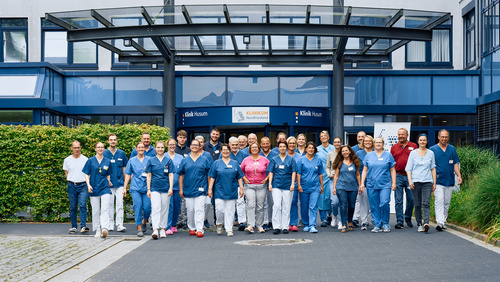 Ein Teil des Teams des Klinikums Nordfriesland befindet sich vor dem Eingang in Husum und kommt dem Betrachter freundlich entgegen.